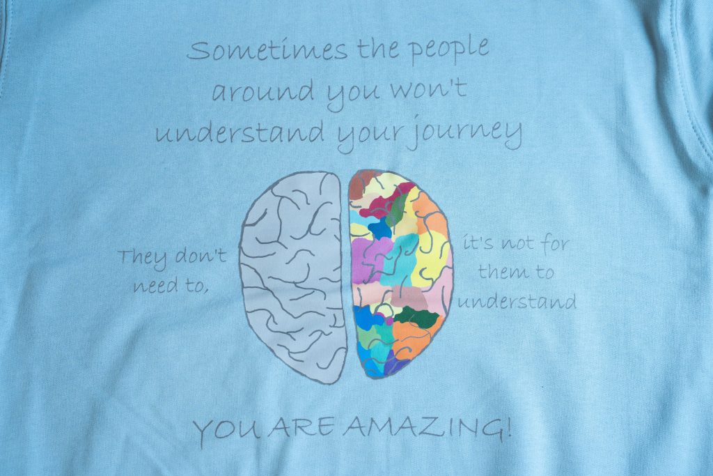 Image of brain on light blue hoodie. Left hemisphere gray, right multicoloured. Text around image states "Sometimes the people around you won't understand your journey. They don't need to, it's not for them to understand. YOU ARE AMAZING!"