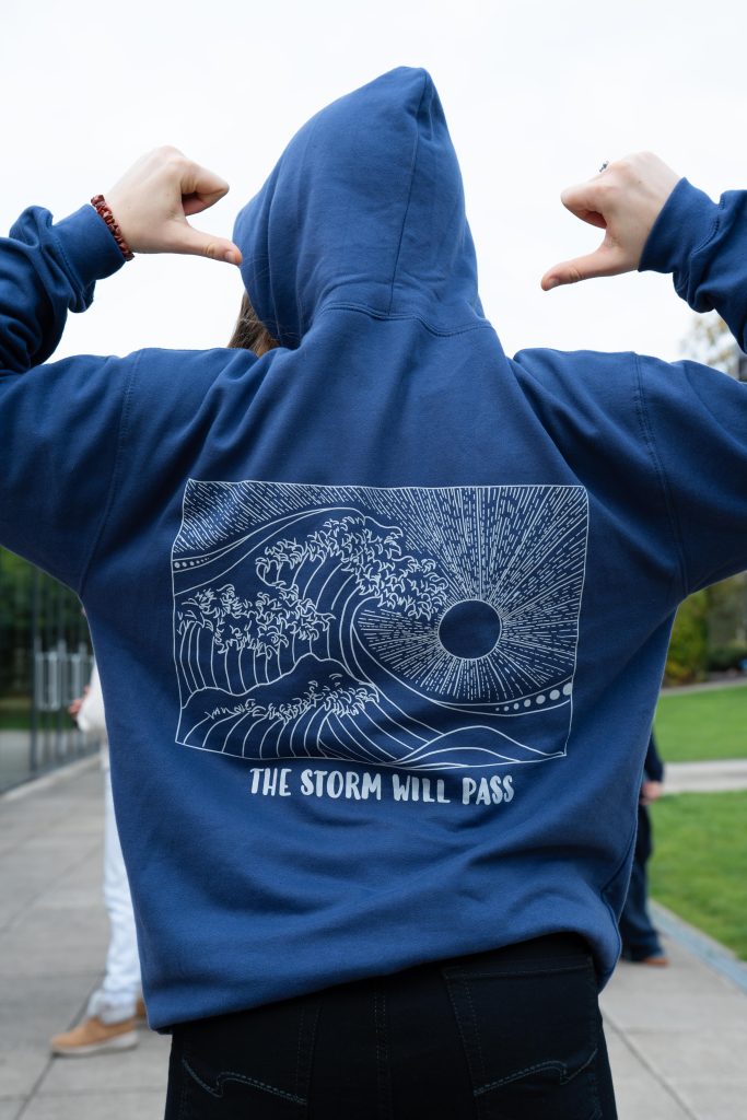 Image of a person with a blue hoodie on, their back facing the camera. There is a line drawing of a sun and waves and underneath it says "The storm will pass"